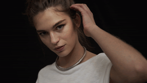 Close-up studio shot of a woman with brown hair and a white t-shirt, wearing a silver necklace against a solid black background.