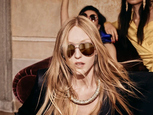 Three women pose indoors, one in sunglasses and a necklace, another in yellow, and another with sunglasses partially visible in background.