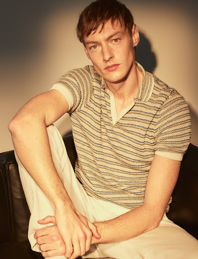 Young male model with styled reddish hair wearing a patterned knit polo shirt and white trousers, seated against a plain wall.