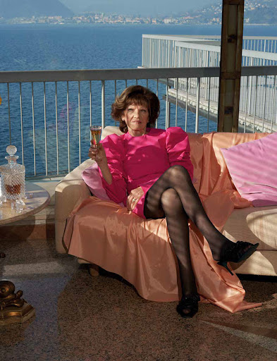 Elegant mature woman in vibrant pink dress, sitting on a couch on a balcony with a sea view, sipping champagne, and wearing black pantyhose.
