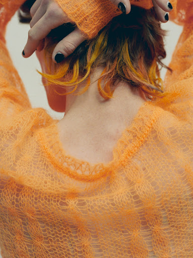 Close-up back view of a person wearing an orange open-knit mohair sweater with hands raised behind the head, featuring black nail polish.