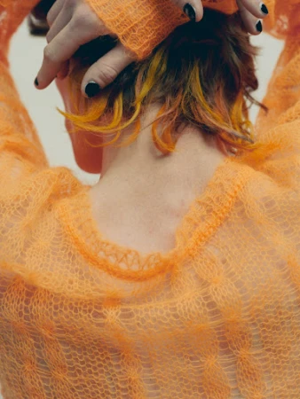 Close-up back view of a person wearing an orange open-knit mohair sweater with hands raised behind the head, featuring black nail polish.
