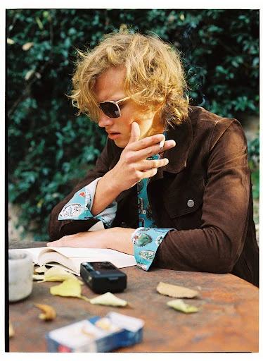 Young adult with blond hair sits outdoors at a table, smoking a cigarette, reading a book with leaves and a pack of cigarettes on table.