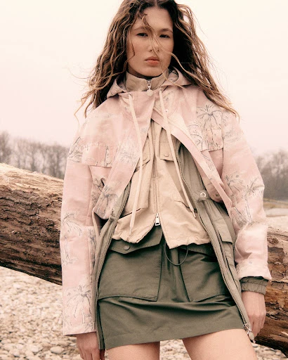 Fashionable woman poses in a scenic outdoor setting wearing a stylish pink and olive green jacket, paired with a matching skirt.