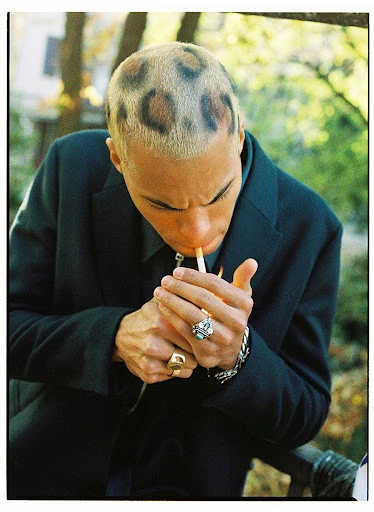A young adult male in a dark suit lights a cigarette, showcasing a striking leopard-print buzzcut in an outdoor setting with soft lighting.