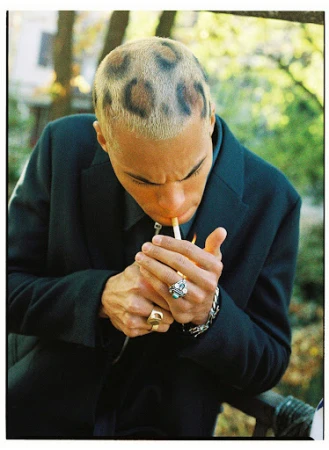 A young adult male in a dark suit lights a cigarette, showcasing a striking leopard-print buzzcut in an outdoor setting with soft lighting.