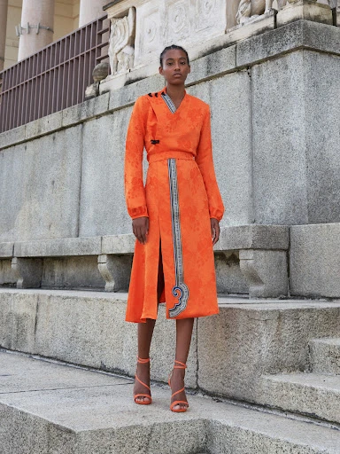 A dark-skinned woman models a knee-length orange wrap dress and strappy heels on a stone staircase with classical architecture.
