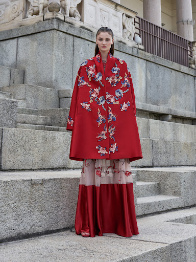 A woman in a floral embroidered red cape and flowing skirt stands on concrete steps of an outdoor structure in a fashion editorial shot.