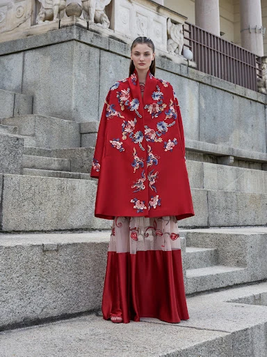 A woman in a floral embroidered red cape and flowing skirt stands on concrete steps of an outdoor structure in a fashion editorial shot.