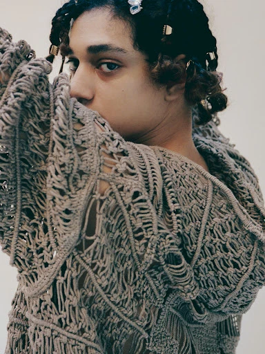 Close-up portrait of a person with braided hair wearing an open-weave knit sweater, with neutral wall background, creating a fashion editorial image.