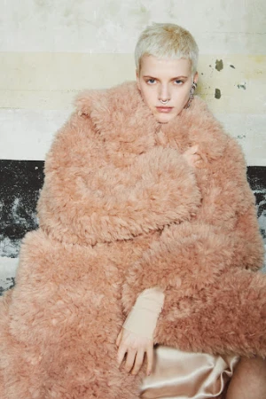 Model with short platinum blonde hair and septum piercing wearing an oversized fluffy pink coat against an aged wall
