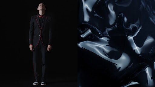 A man in a dark suit stands against a black background next to an abstract rippled black surface with white highlights in the studio.