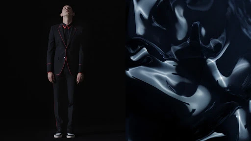 A man in a dark suit stands against a black background next to an abstract rippled black surface with white highlights in the studio.