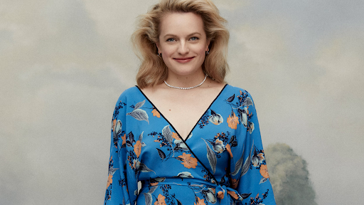 Smiling woman with blond hair poses in a blue floral wrap dress and necklace against a soft-focus studio background.