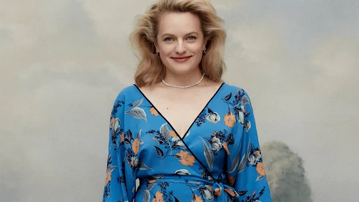 Smiling woman with blond hair poses in a blue floral wrap dress and necklace against a soft-focus studio background.