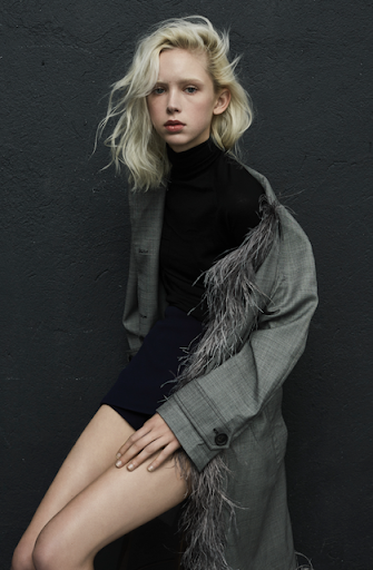 Young woman with platinum blonde hair poses in a grey coat with feather trim against a dark grey textured wall in a studio fashion shot.