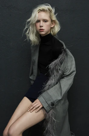 Young woman with platinum blonde hair poses in a grey coat with feather trim against a dark grey textured wall in a studio fashion shot.