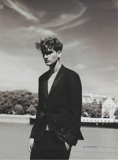 Monochrome portrait of a young man with styled hair wearing a dark blazer outdoors by water under a cloudy sky.