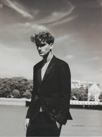 Monochrome portrait of a young man with styled hair wearing a dark blazer outdoors by water under a cloudy sky.