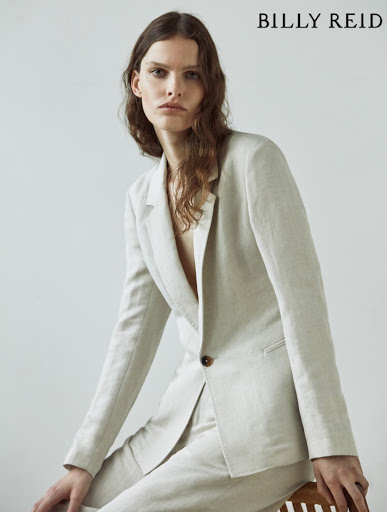 Portrait of a woman in a light-colored linen suit, seated on a wooden stool, gazing directly at the camera with a neutral expression.