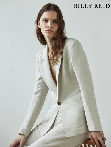 Portrait of a woman in a light-colored linen suit, seated on a wooden stool, gazing directly at the camera with a neutral expression.