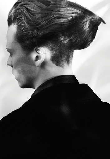 Monochrome portrait of an adult male with a distinctive hairstyle wearing a dark jacket against a light background, shot in profile.