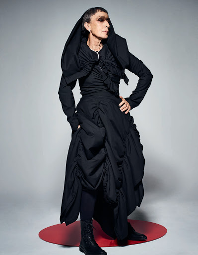 Woman in dramatic black avant-garde fashion, including a draped jacket and voluminous skirt, stands in a studio setting with soft lighting.