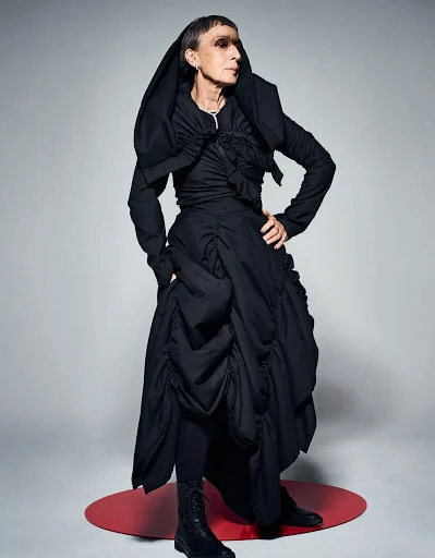 Woman in dramatic black avant-garde fashion, including a draped jacket and voluminous skirt, stands in a studio setting with soft lighting.