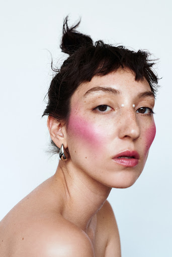 Close-up portrait of an adult woman with fair skin, dark hair, and pink makeup, in front of a simple white background in a studio.