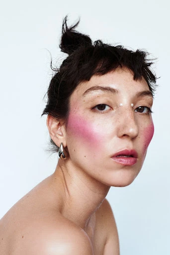 Close-up portrait of an adult woman with fair skin, dark hair, and pink makeup, in front of a simple white background in a studio.