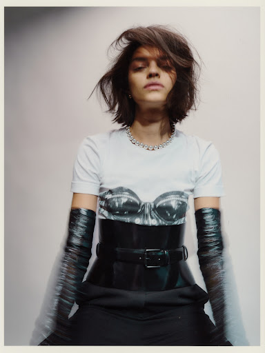 Portrait of a woman with short hair wearing a graphic t-shirt, corset belt, and long gloves against a gradient grey background.