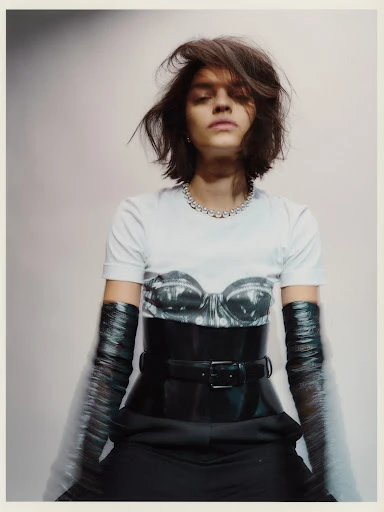 Portrait of a woman with short hair wearing a graphic t-shirt, corset belt, and long gloves against a gradient grey background.