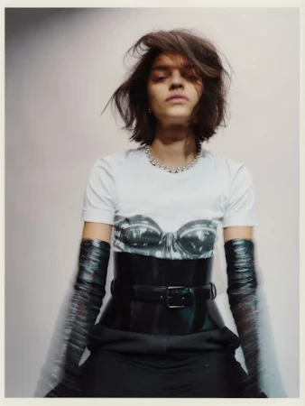 Portrait of a woman with short hair wearing a graphic t-shirt, corset belt, and long gloves against a gradient grey background.