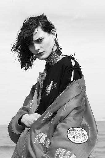 Portrait of a stylish woman in a bomber jacket with the word 'FEAR' printed on it, posing outdoors with a neutral expression against a sky backdrop.