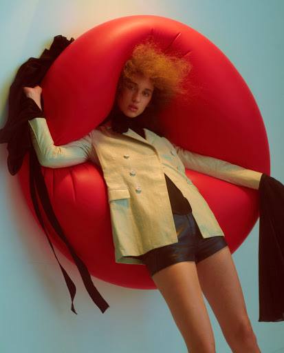 Model with voluminous curly hair posed inside a giant inflatable red circle wearing a pale yellow blazer and black shorts
