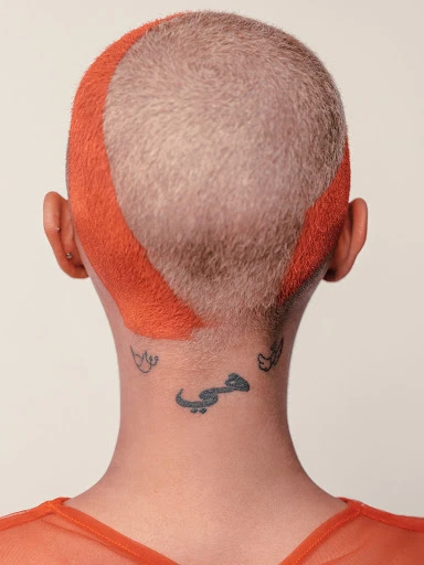 Back of a person's head with an orange-dyed buzz cut and multiple tattoos on the neck. Focus on personal style and body art.