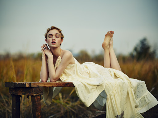Young woman in a yellow dress lies on a wooden bench outdoors in a field with tall brown grass, legs raised, looking at the camera.