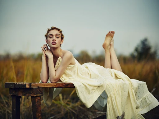 Young woman in a yellow dress lies on a wooden bench outdoors in a field with tall brown grass, legs raised, looking at the camera.