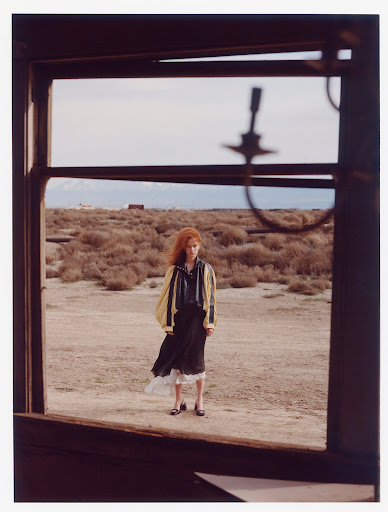 Full-length portrait of a woman in a yellow jacket and black dress standing in a desert landscape framed by a broken window.