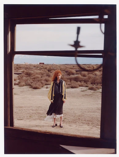 Full-length portrait of a woman in a yellow jacket and black dress standing in a desert landscape framed by a broken window.