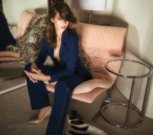 A woman sits in a pale pink chair wearing a dark blue suit in an indoor setting with a side table and decorative carpet visible.