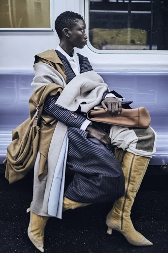 Fashionable person in a pinstripe suit with a tan coat and boots, sitting in a subway car with a neutral expression.
