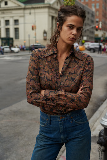 A young woman with long brown hair and a patterned shirt stands outdoors on a city street with a serious expression on her face.