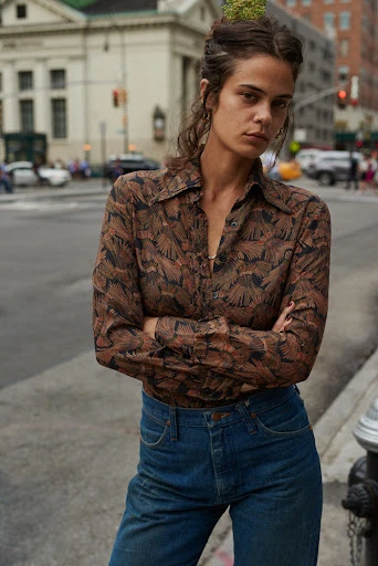 A young woman with long brown hair and a patterned shirt stands outdoors on a city street with a serious expression on her face.