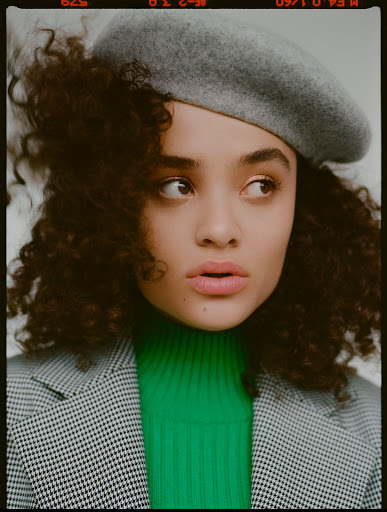Portrait of a young woman with curly hair wearing a grey beret, green turtleneck sweater, and blazer looking off to the side.