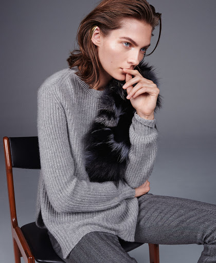 Adult male fashion model poses in studio, wearing a grey sweater, grey trousers, and a fur collar on a chair against grey background.