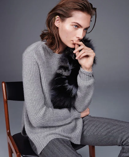 Adult male fashion model poses in studio, wearing a grey sweater, grey trousers, and a fur collar on a chair against grey background.