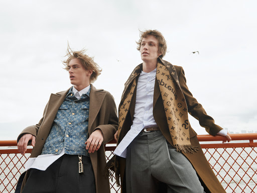 Two adult males pose outdoors wearing brown coats and patterned shirts and scarves, against a neutral sky and fence.