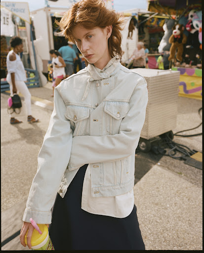 A person with red hair stands in front of an outdoor fair with muted tones wearing a denim jacket and holding a yellow drink.
