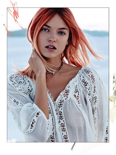 Woman with red hair wears an off-white boho blouse in front of a blurred pale blue sea, posing in a medium shot.